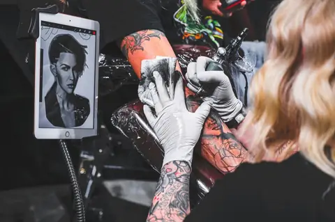 Artist tattooing a colour sleeve from a reference portrait on a tablet